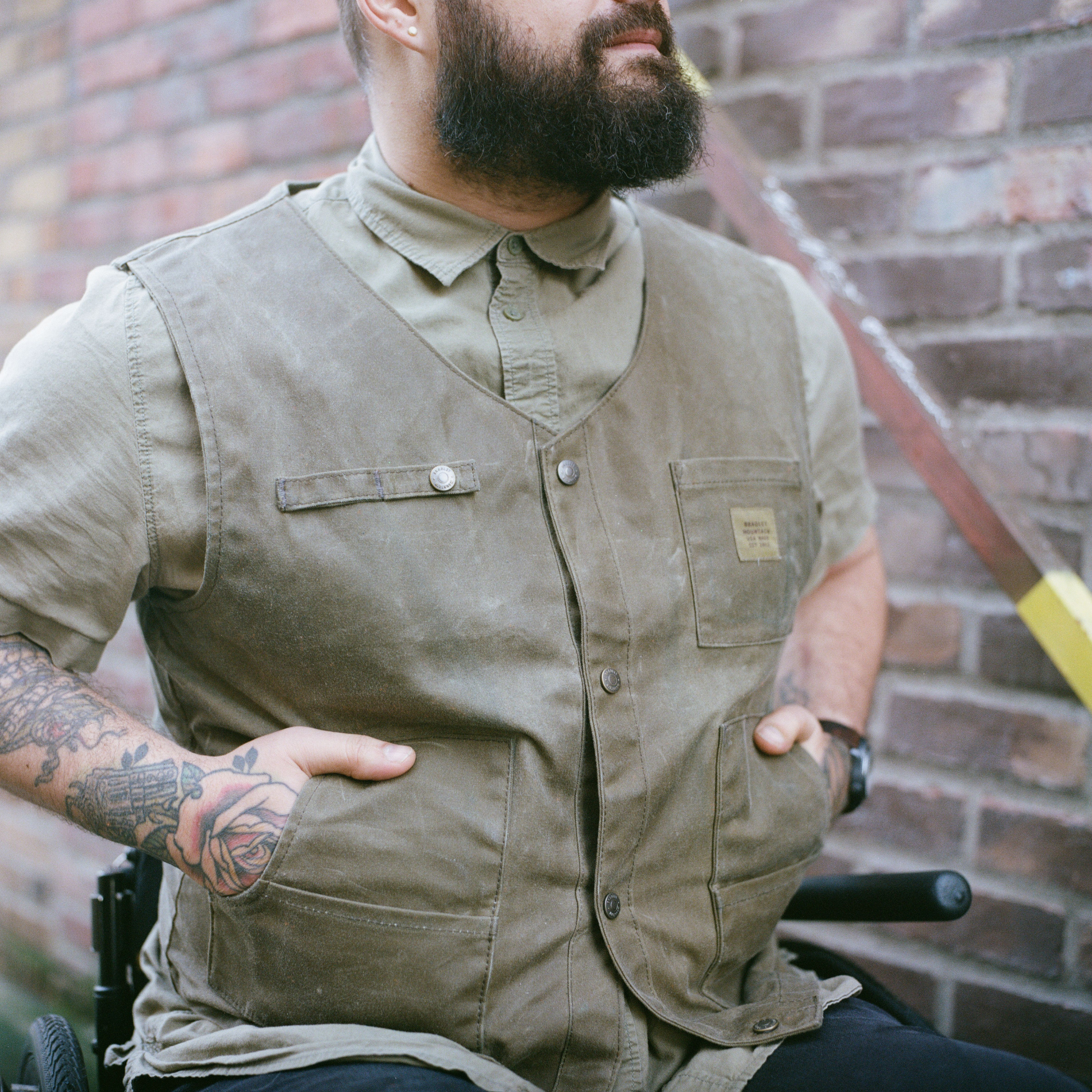 An image of a person wearing a waxed canvas Bradley Mountain Nomad Vest in Field Tan with hands in pockets. The hands have tattoos.