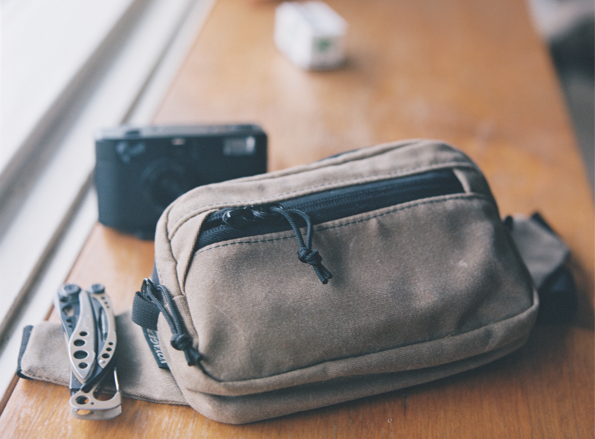 An image of a small handmade waxed canvas sling bag placed on a wooden surface with a pocket knife, and a camera in the background.