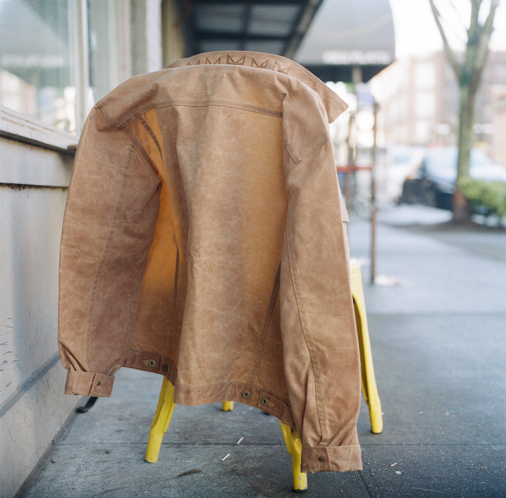 A Brown Ginew waxed canvas Rider jacket on a yellow chair outdoors in front of Porchlight Coffee.