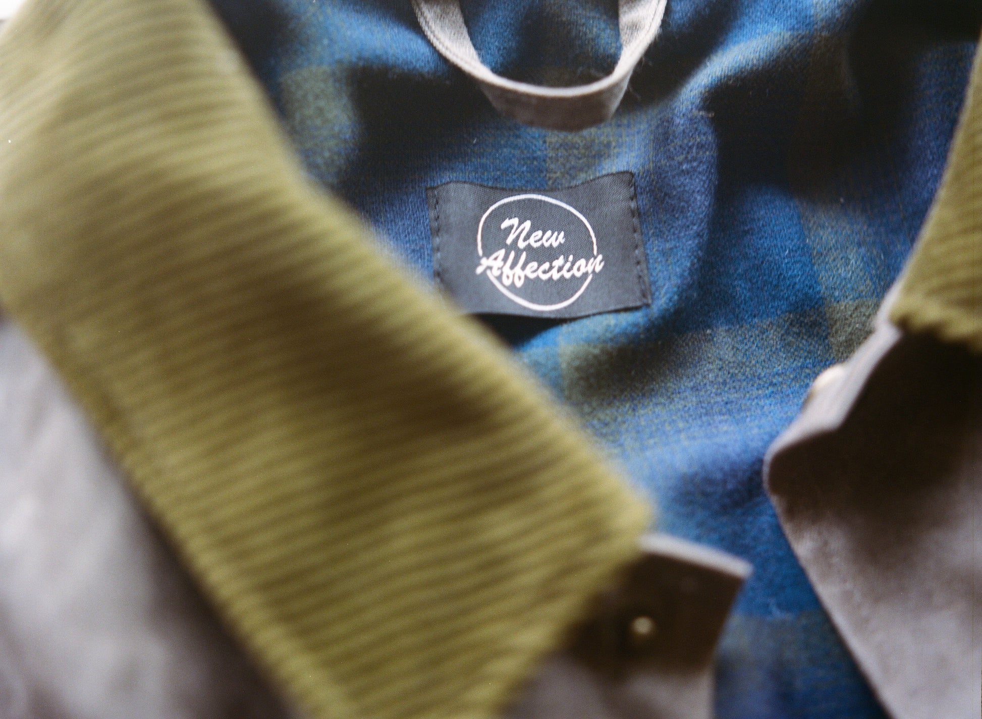 A black label with white writing that says "New Affection" on green and blue plaid background. A green corduroy collar is blurry in the foreground.