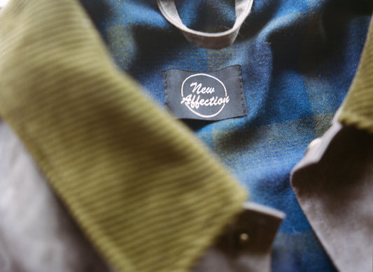 A black label with white writing that says "New Affection" on green and blue plaid background. A green corduroy collar is blurry in the foreground.