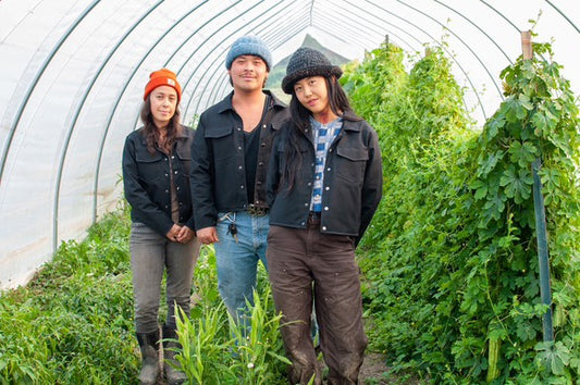 Three people with dark blue waxed jackets standing in a greenhouse together with plants around them.