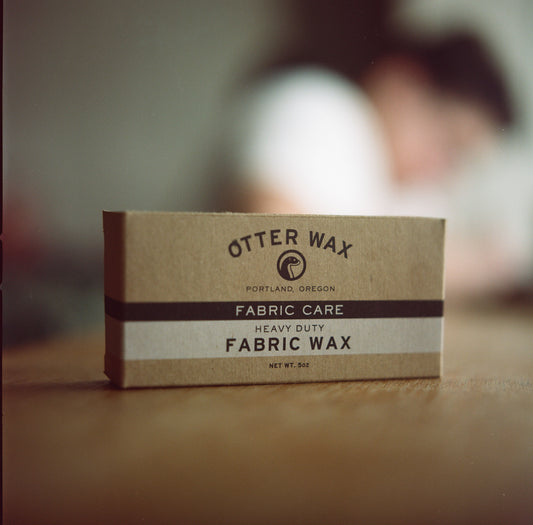 A small cardboard box that says "Otter Wax Heavy Duty Fabric Wax," "Made in Portland, and "5 ounces" on a wood surface. A person wearing a white t shirt is blurred in the background.