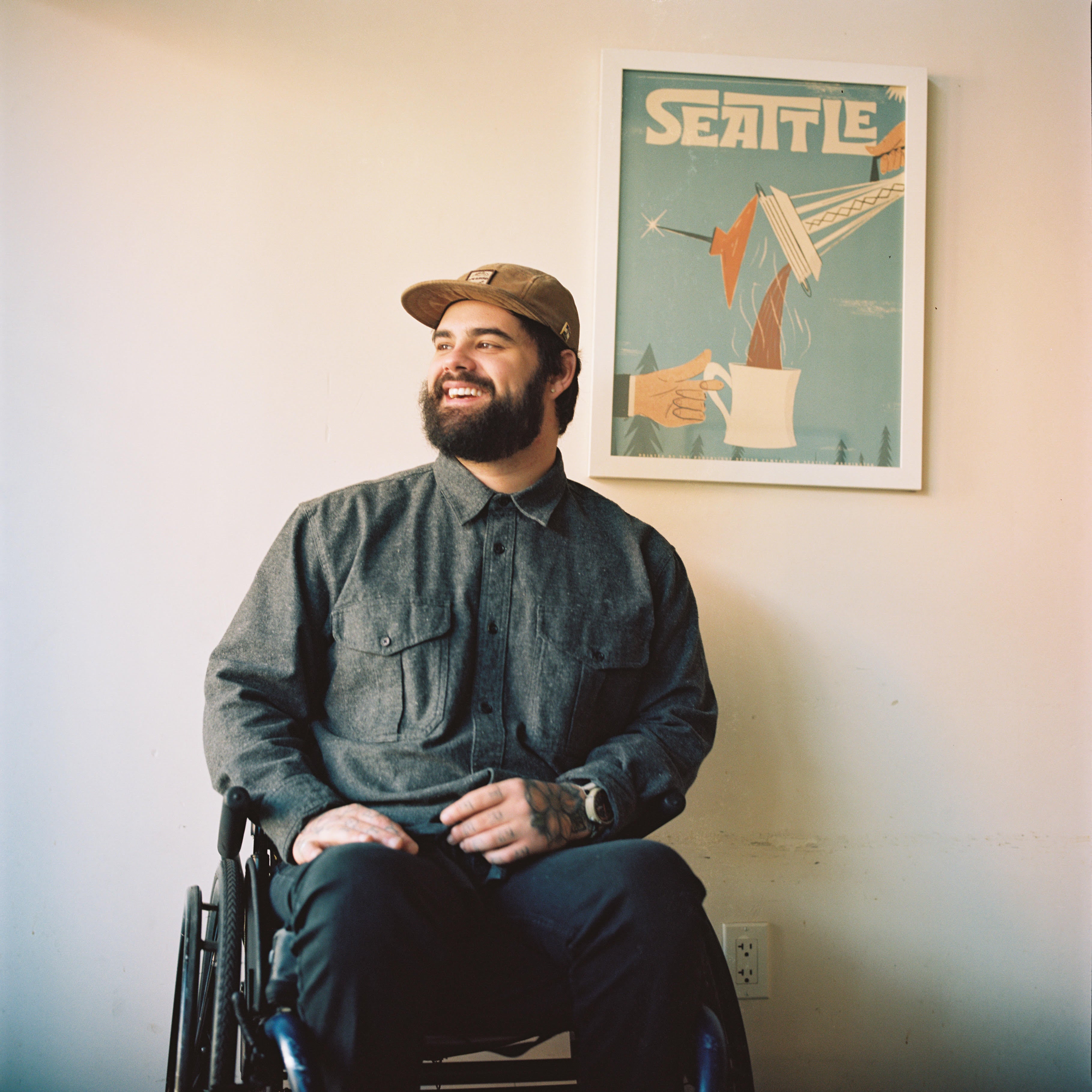 An image of Weathered Waxing owner Cameron in his wheelchair in front of a white wall with a Seattle poster made by Porchlight Design Co hanging in the background..